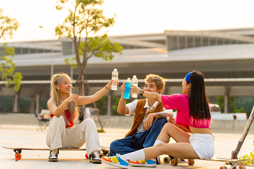 Group of Asian man and woman friends resting and drinking water together after longboard skating at public park. Stylish youth people enjoy urban outdoor lifestyle extreme sports skateboarding on summer vacation.