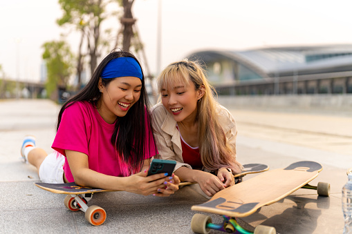 Group of Stylish youth people Asian man and woman friends have fun urban outdoor lifestyle using mobile phone with internet during skating on longboard skate at city street together on summer vacation.