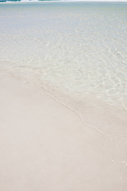 This color image if of beach front property with a beautiful view of the ocean. the picture includes a deck with deck chair next to the beach front with sand and ocean. this picture is a classic scenic beach front setting that is beautiful. there are also waves crashing against the beach. the mood is very peaceful and relaxing. the lighting is natural sunlight. and the background consists of the blue ocean and blue sky. 