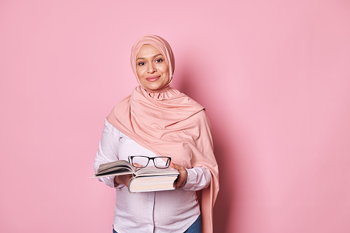 Confident multi-ethnic middle aged Muslim woman educator, in pink hijab, a school teacher holding eyeglasses and studying book, looking at camera, isolated on pink background. World Teacher's Day