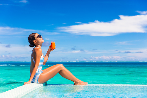Summer healthy eating woman drinking detox carrot juice for balanced diet weight loss bikini body lifestyle. Happy living girl enjoying sun tan holidays by the swimming pool of tropical hotel.