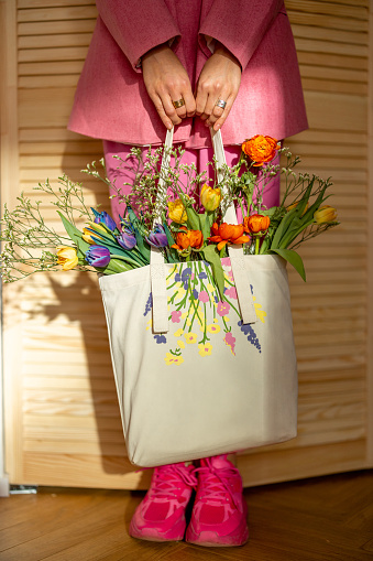 Stylish woman in pink holds totebag filled with flowers indoors, close-up on bag. Spring, style and eco-bag concept