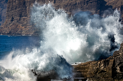 The mighty ocean sends a massive wave crashing against the rocky shore, creating a spectacular display of natural power, as the Acantilados de los Gigantes cliffs tower in the background of this scene