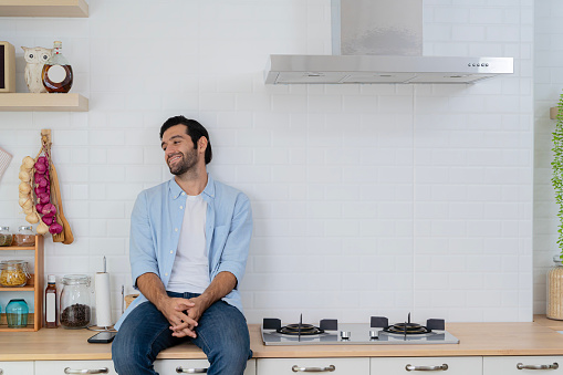 Attractive man sitting in a home kitchen. A handsome young man looks out the window as he sits on the kitchen counter.