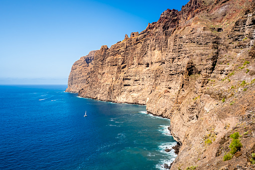 Los Gigantes cliffs tower above the rocky coastline of Tenerife's western shore while a small sailboat sails through the calm waters, creating a striking contrast for nature enthusiasts vacations.