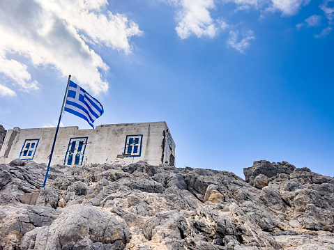 Travel destination concept: Greek flag waving in the wind, hanging on a pole, over a rock at the coastside at the edge of an island. View from below on building, blue sky and white clouds. Nature background with copy space.