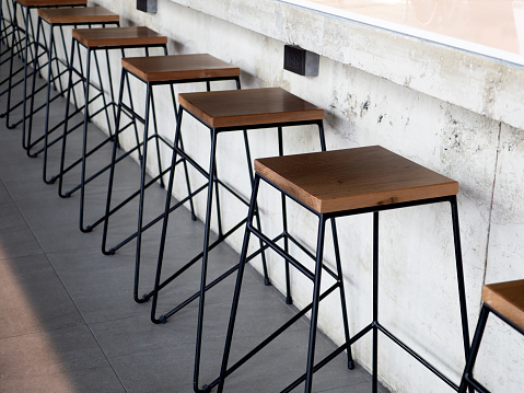 Row of wooden bar stool chairs beside concrete counter bar, loft style cafe interior. Empty wood seats with black steel bar decoration on concrete floor with copy space.