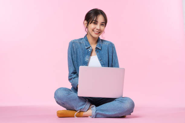 istock Young Asian college student sits cross-legged on floor working with a laptop computer, looking directly at the camera in a studio shot on a pink background 1482589585