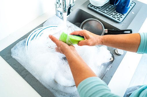 High angle view of woman's hands washing a drinking glass with a sponge in a kitchen sink. High resolution 42Mp indoors digital capture taken with SONY A7rII and Zeiss Batis 25mm F2.0 lens