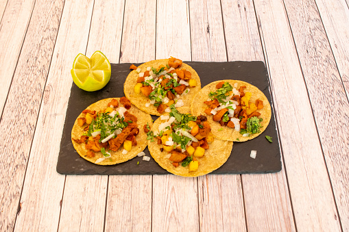 Tacos al pastor in corn tortillas filled with pineapple, meat, cilantro and chopped white onion with a chopped lime on black slate