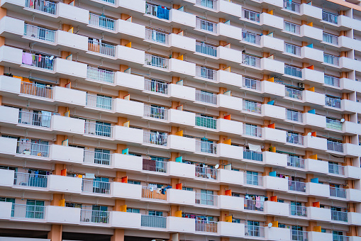The exterior of a Japanese apartment complex with white walls and railings. The building features numerous balconies, each with its unique set of items displayed.