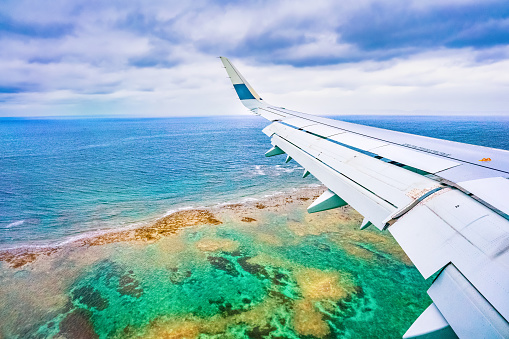 An airplane as it approached Naha Airport in Okinawa. The view outside the window shows the airplane wing and below it, a stunningly beautiful coastline with crystal-clear blue-green waters and coral reefs.