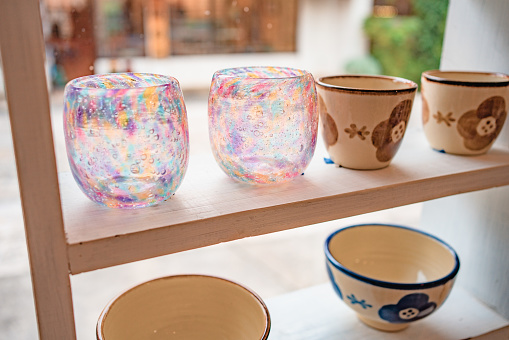 Various handmade cups and bowls are displayed on shelves in an Okinawan pottery shop, including two pink and transparent glass cups.