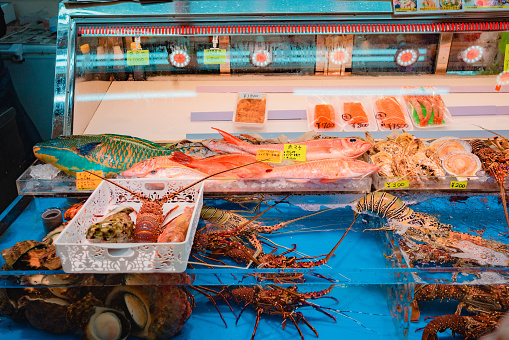 A seafood vendor in the Makishi Market in Okinawa, Japan. The vendor has a water tank with live lobsters and a shelf with various fresh seafood such as fish, shellfish, and sashimi.