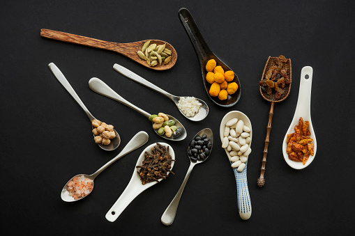 Set of messy spoons of different materials and shapes filled with nuts, seeds, spices and legumes on black background