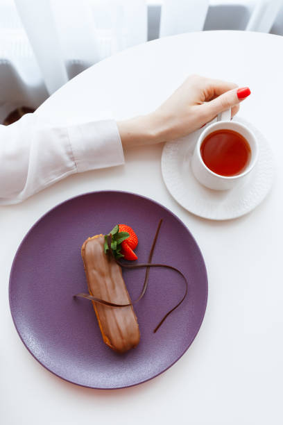 白いテーブルの上に紅茶を飲んだ女性の手の接写。新鮮なイチゴとチョコレートエクレアの紫色のプレートの近く。5時のお茶。 - five oclock tea ストックフォトと画像