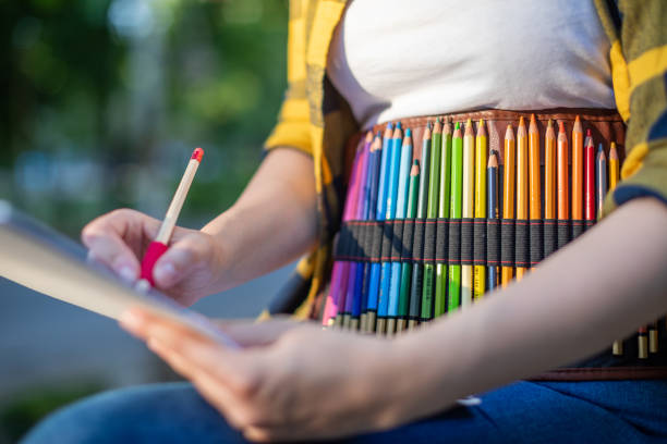 A young woman is sitting outside and holding a sketching pad in her hands. She has a pencil case around her waist and she is drawing. Unrecognizable person.