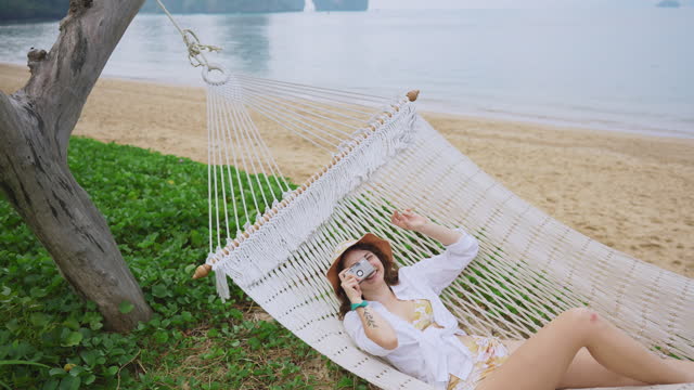 Woman laying in hammock and photographing with camera on the background of the sea