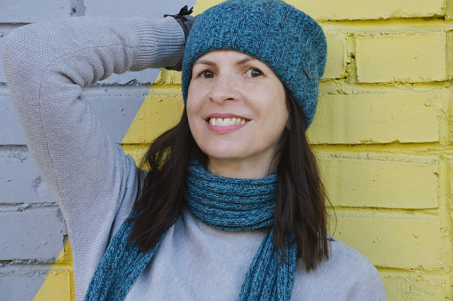 A young woman in a gray sweater and a turquoise hat and scarf stands near a colored brick wall. Urban portrait of a girl.