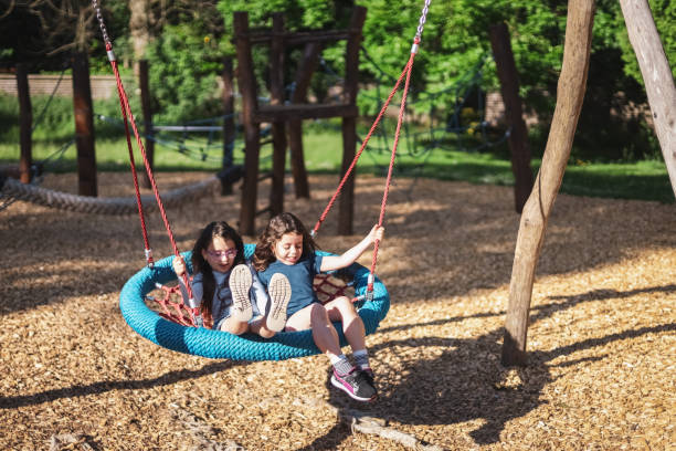 Two caucasian girls flying at speed with hair fluttering in the wind sitting on a round rope Portrait of two beautiful caucasian girls flying at speed with smiles and joyful real emotions, sitting on a round rope swing in the park at the playground, close-up side view. The concept of PARKS and SECs, happy childhood, children picnic, holidays, children recreation, outdoors, playgrounds, children entertainment. funny camping signs pictures stock pictures, royalty-free photos & images