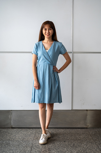 Full length image of smiling Asian woman wearing blue turquoise dress with wall background