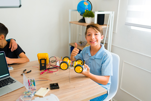 Excited successful teen giving a thumbs up while finishing his electronic robot prototype during a junior high class