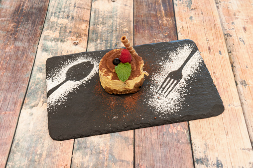 Tiramisu cake with berries, cocoa powder, icing sugar and biscuit wafer on a black slate plate