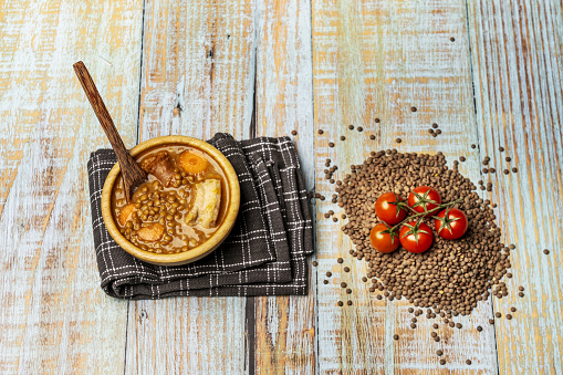Still life with traditional lentil stew in a wooden bowl on a light wooden table with dried lentils and strings of cherry tomatoes