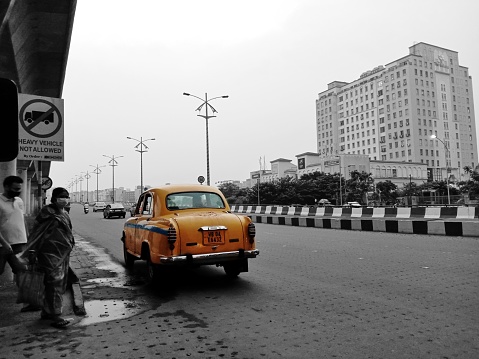 This editorial photograph captures a slice of life in the bustling city of Kolkata, specifically in the rapidly developing area of New Town Rajarhat. In the foreground of the photograph, a group of people are seen waiting for the bus near the New Town bridge, while in the background, the iconic yellow taxi of Kolkata is parked by the roadside. The photograph has been rendered in monochrome, except for the vibrant yellow of the taxi, which stands out prominently in the frame. The composition of the photograph highlights the coexistence of old and new elements in the city, with the classic yellow taxi juxtaposed against the sleek modern architecture of the surrounding buildings. Overall, this photograph captures the essence of Kolkata's unique blend of tradition and modernity.