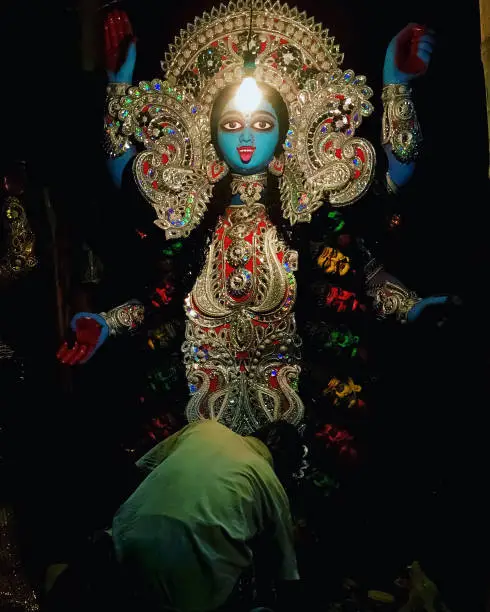 This photograph captures the essence of Kali Puja, a festival celebrated in West Bengal, India. The photo was taken at Kumartuli, where artisans make clay idols of the goddess Kali. The artist seen in the photo is giving the finishing touch to the idol before the festival. The vibrant darkness of the surroundings is illuminated by a filament bulb placed on the forehead of the goddess, creating a mesmerizing effect. The image showcases the beauty of the festival and the artistry involved in its celebration.