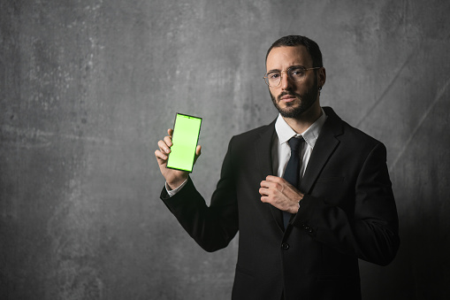 Portrait of a handsome businessman in suit standing in front of the wall in studio, he is holding a smart phone with green screen.