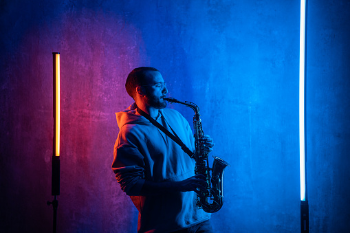 Man playing saxophone in dark studio with neon lighting on the wall.