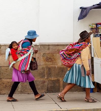 Sucre, Bolivia, April 1 2023 : two Bolivian women walking in the street with colored traditional clothes and hats. One of them is carrying a baby behind her back.