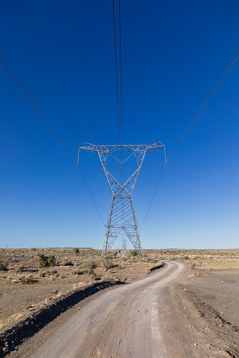 High voltage electricity transmission pylons and lines with a gravel road next to it in the Karoo area of South Africa