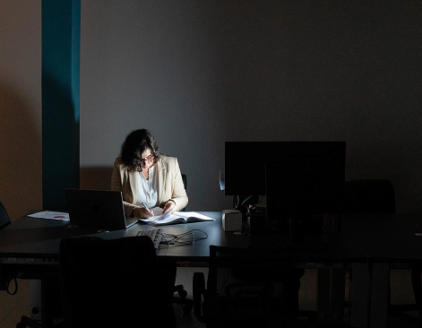 Mature businesswoman illuminated by a lamp while writing notes in a file at late at a desk in a dark office