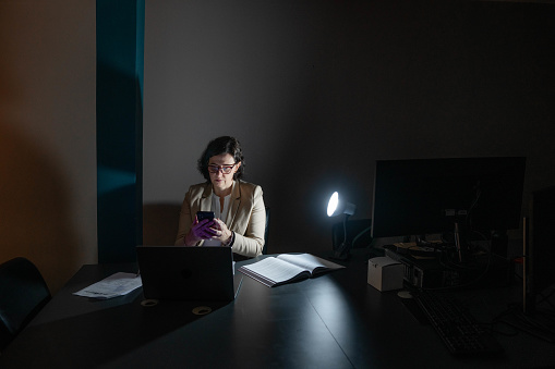 Mature businesswoman illuminated by a lamp texting on her phone at a desk while working late in a dark office