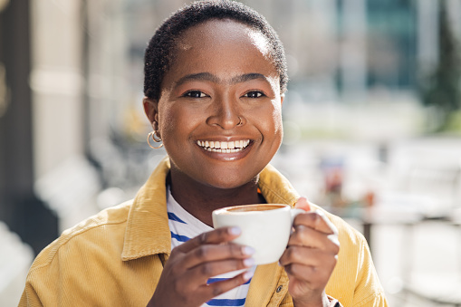 Headshot of an African American woman drinking a coffee in an open-air cafeteria in the city center. She is glowing!