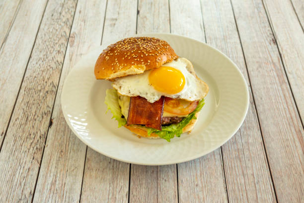 istock Classic beef burger with fried egg to dip the yolk and fried bacon with melted cheese, lettuce and ketchup on a white plate 1482666055