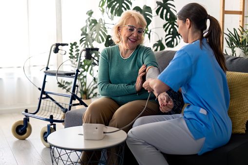 Beautiful senior woman measuring blood pressure with help of home nurse, sitting on sofa in living room, feeling satisfied and happy with home care. Female nurse assisting senior woman to measure blood pressure