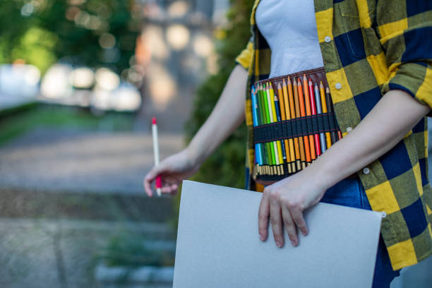 A young woman holding a sketching pad in her hands. She has a pencil case around her waist and she is walking. Unrecognizable person.