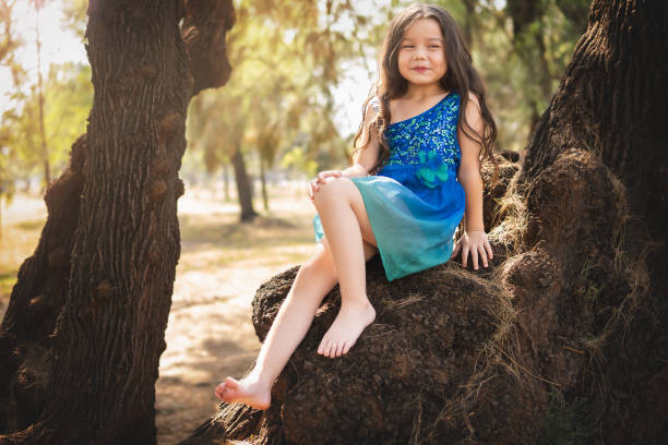 beautiful happy girl playing in the park with blue dress without shoes sitting on tree laughing screaming with happiness in family enjoying children's day stock photo