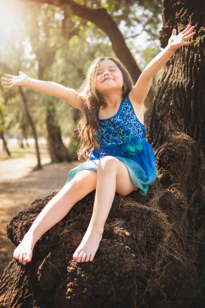 beautiful happy girl playing in the park with blue dress without shoes sitting on tree laughing screaming with happiness in family enjoying children's day stock photo
