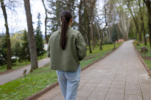 Rear view of young casually clothed woman slowly walking through alley of green trees on sidewalk at city park