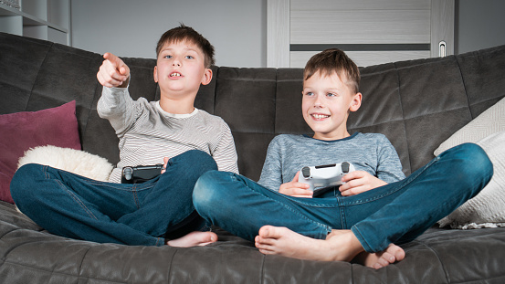 Happy teenagers sit on sofa with game joysticks. Children play videogames in home room. Boy pointing finger ahead. Interesting amusement, hobby, electronic gaming. Best friends lifestyle.