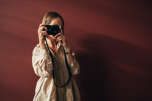 A portrait of a  smiling Caucasian photographer holding her camera while standing in her studio and taking photos. (isolated on red background, copy space)