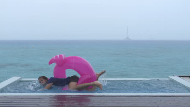 Funny fail video in vacation rain of holidays getaway travel raining away under heavy rainfall, with man falling while trying to get out of flamingo float in luxury pool with funny face.