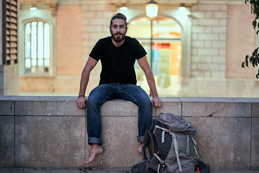 bearded caucasian young man sitting on a wall next to a large backpack smiling at camera barefoot - Backpacker travel concept