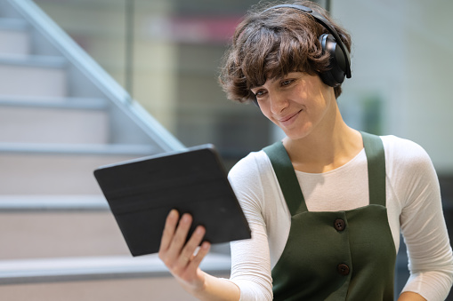 Smiling girl sitting on the stairs of modern architecture, holding a tablet in her hands, cheerfully talking to someone online through the tablet, laughing, listening to music through wireless headphones, looking at the camera. Copy space.