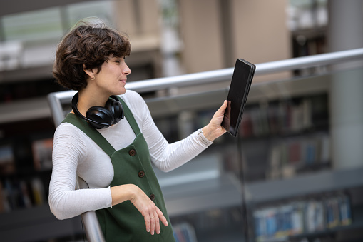 A young cute girl is standing on the top of the stairs with wifi headphones around her neck, holding a tablet, smiling and looking at someone, looking at the camera. Copy space.