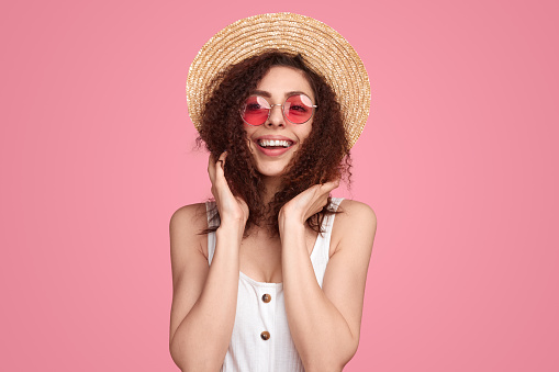 Positive female in sunhat and stylish sunglasses touching curly hair and looking away on pink background in studio